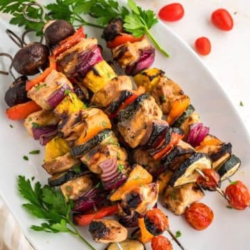 Five full shish kabobs with chicken and colorful vegetables laying diagonally next to a bowl of rice.