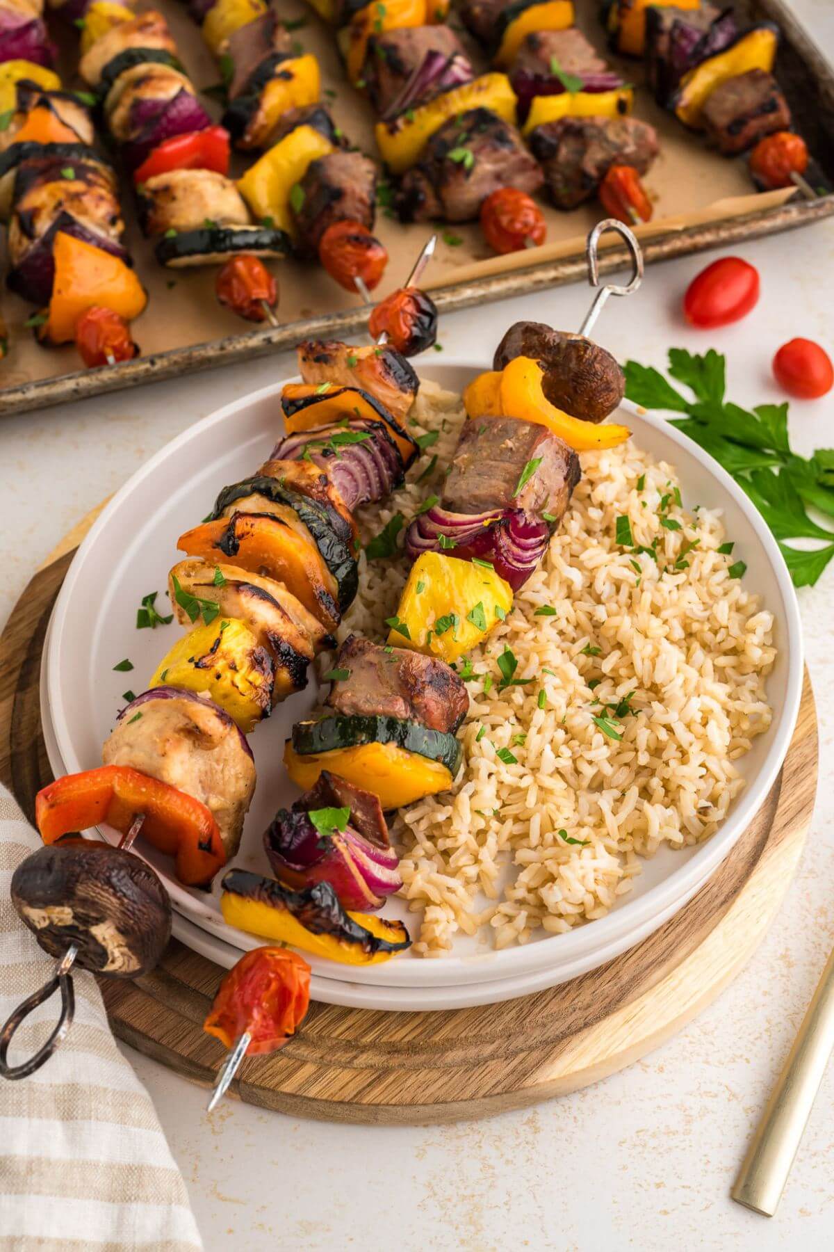 Two completed shish kababs with marinated steak and chicken on a bed of rice next to sheet pan full of assembled kababs.