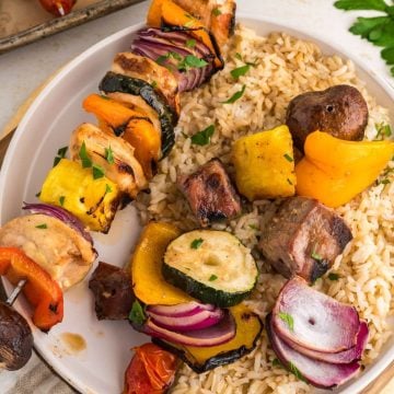 Shish kabab both steak and chicken on a plate over a bed of rice with grilled veggies and pineapple on plate.