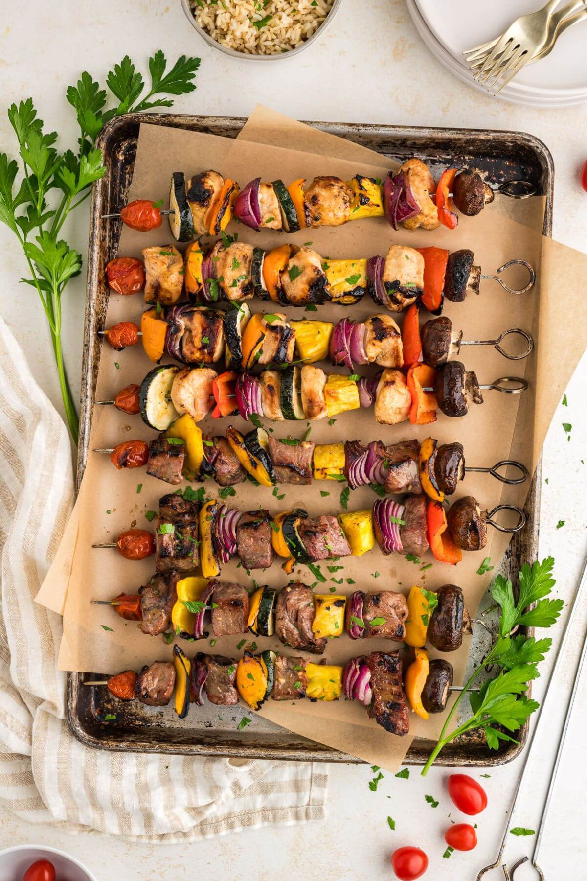 Sheet pan with eight shish kabobs assembled horizontally, set up on table next to napkin, parsley, and dishes