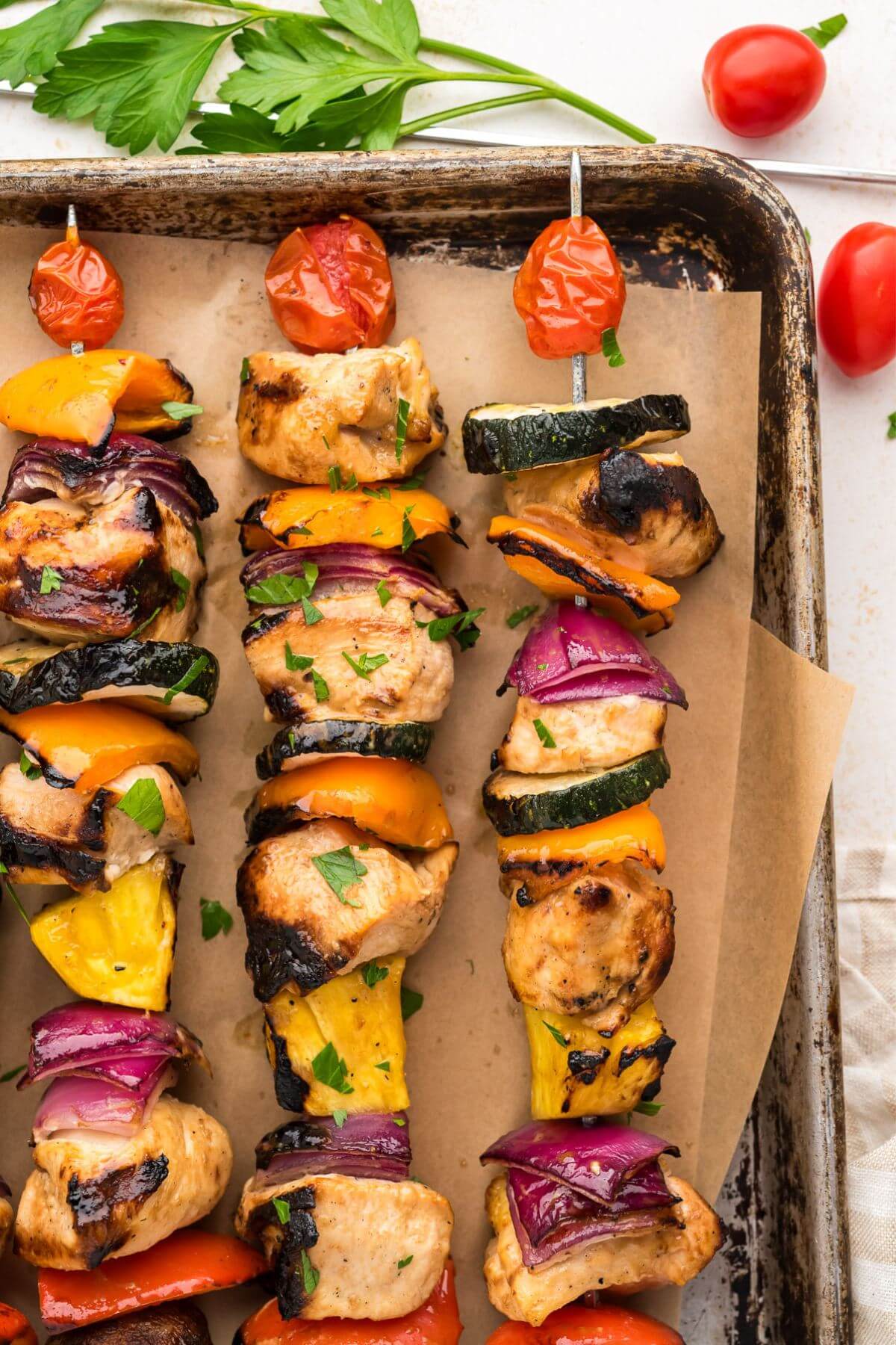 Three shish kabobs assembled vertically on a sheet pan bordered by small tomatoes.