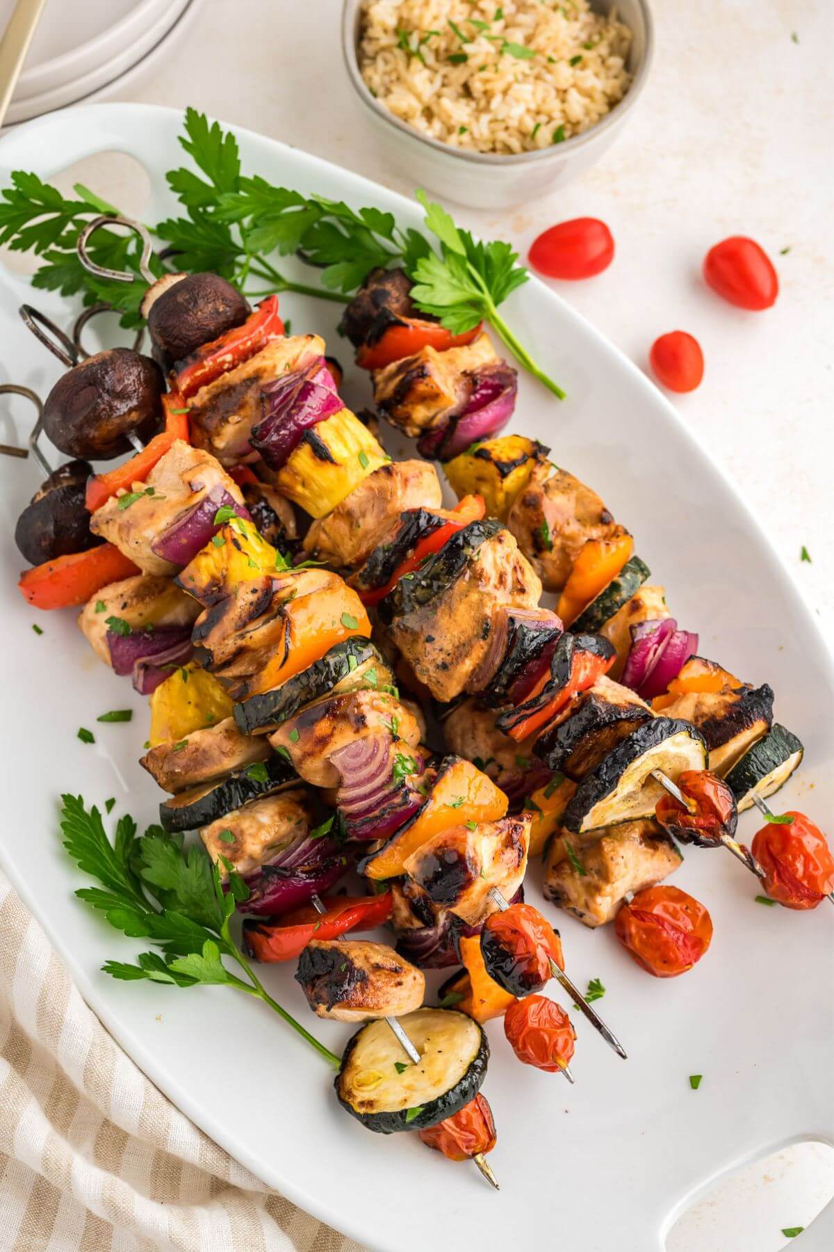 Five full shish kabobs with chicken and colorful vegetables laying diagonally next to a bowl of rice.