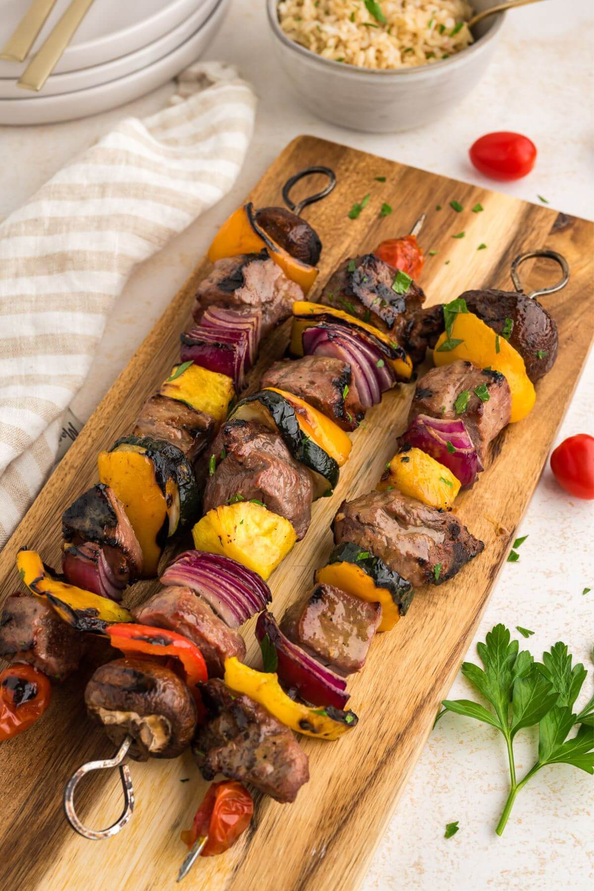 Three assembled shish kabobs with steak and colorful vegetables on a cutting board next to napkin and bowl of rice.