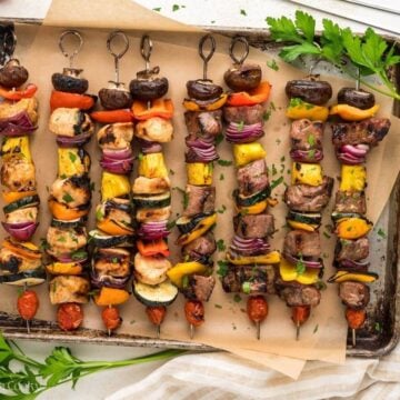 Shish kabobs both chicken and steak grilled and with veggies on a parchment lined tray.