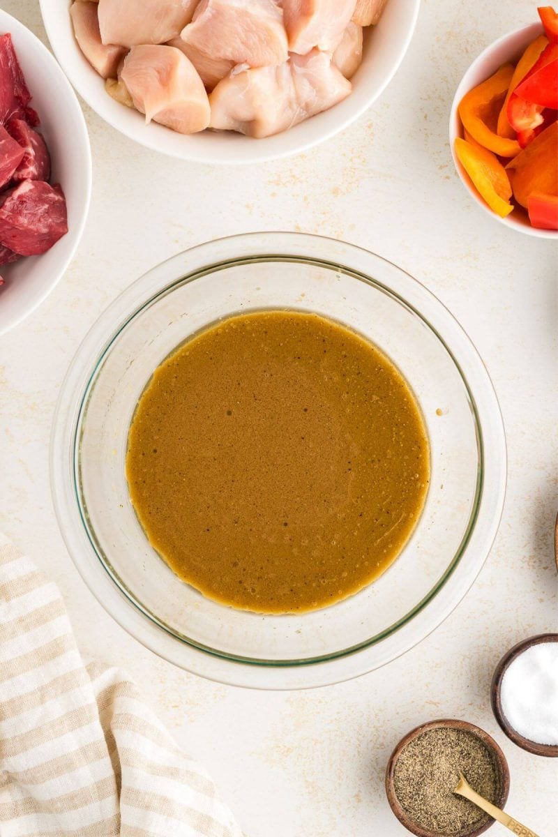 A bowl of the marinade mixture for the shish kababs surrounded by the uncooked kabab meat and vegetables.