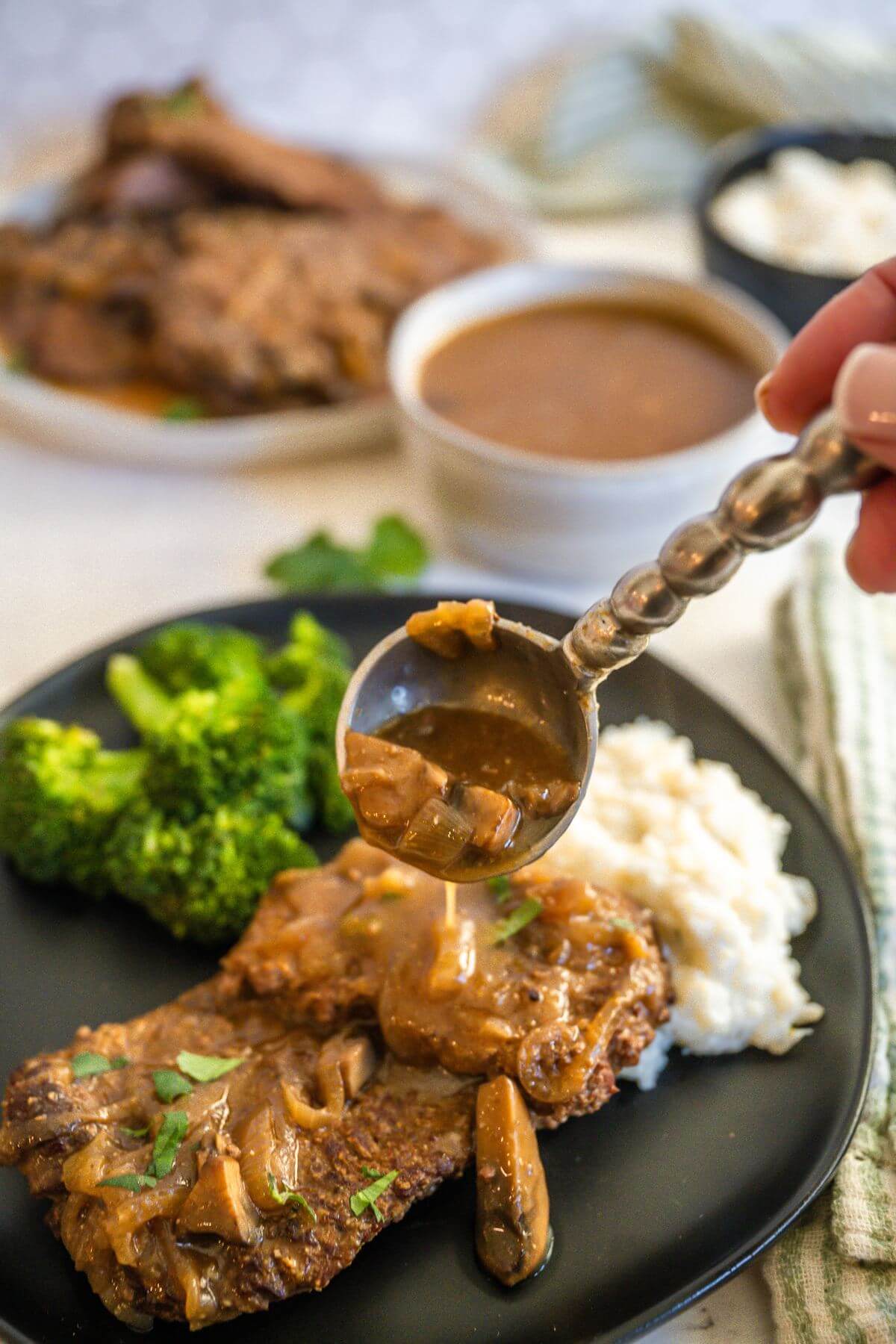 Someone's hand uses fancy spoon to pour gravy on cube steak on plate.
