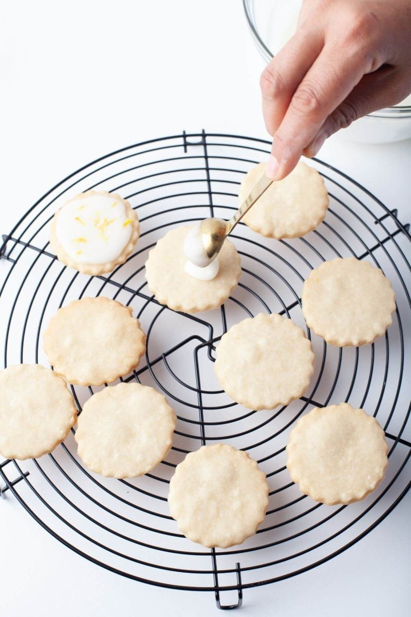 Someon's hand is using spoon to drizzle icing onto cookies on wire rack.