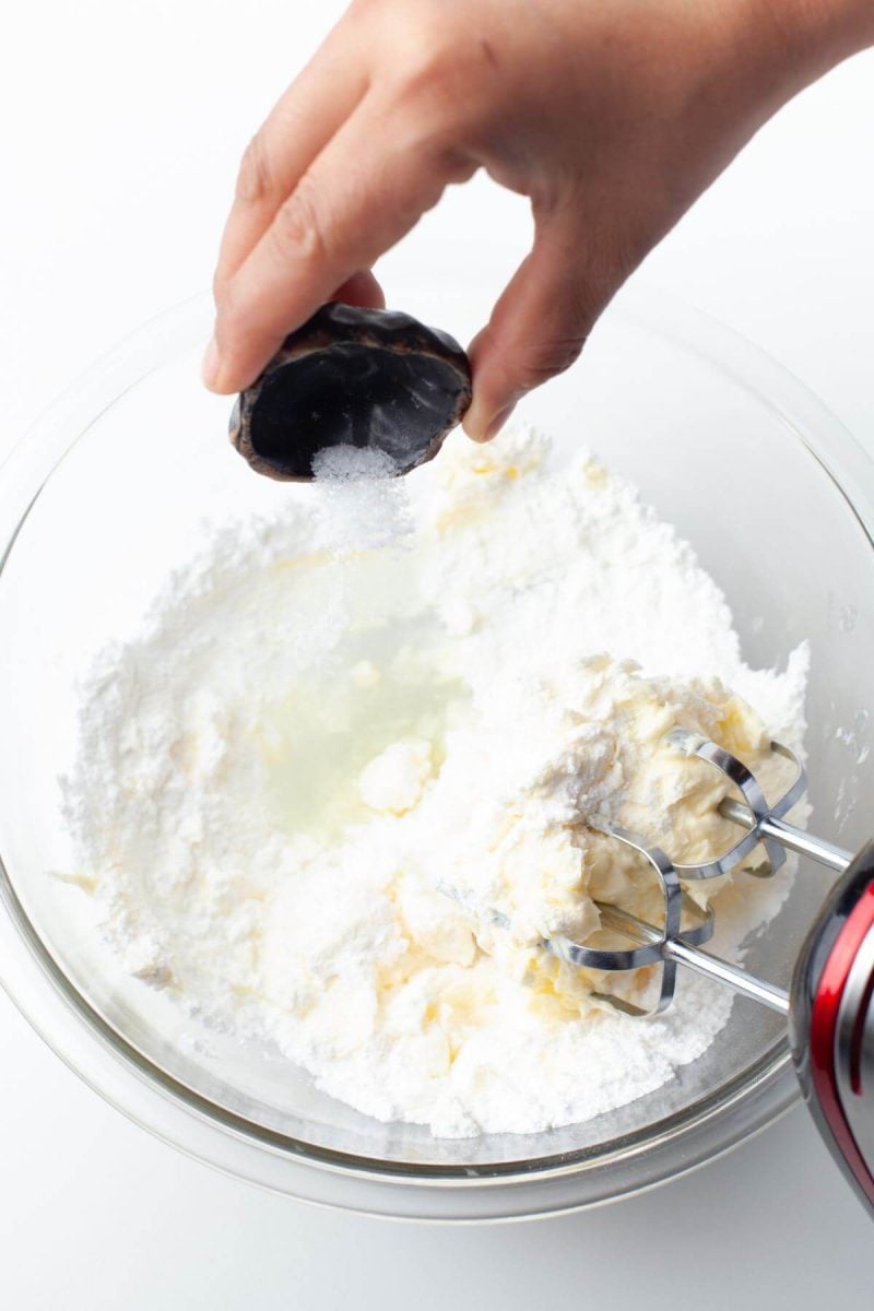 A hand pours tiny portion of salt into mixing bowl filled with flour and creamed butter.