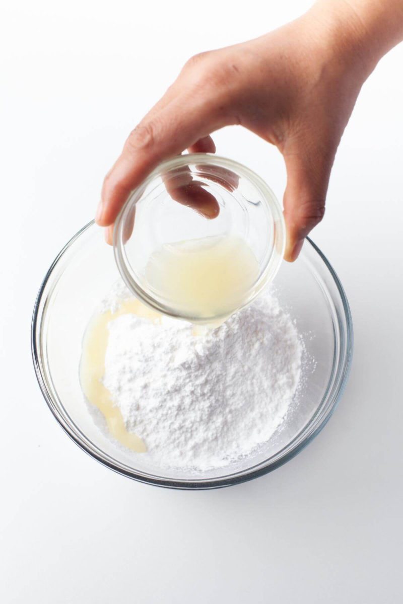 A hand pours lemon juice into bowl of powdered sugar.