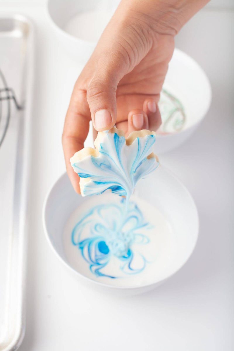 A hand holds a blue marbled leaf cookie over the icing bowl.