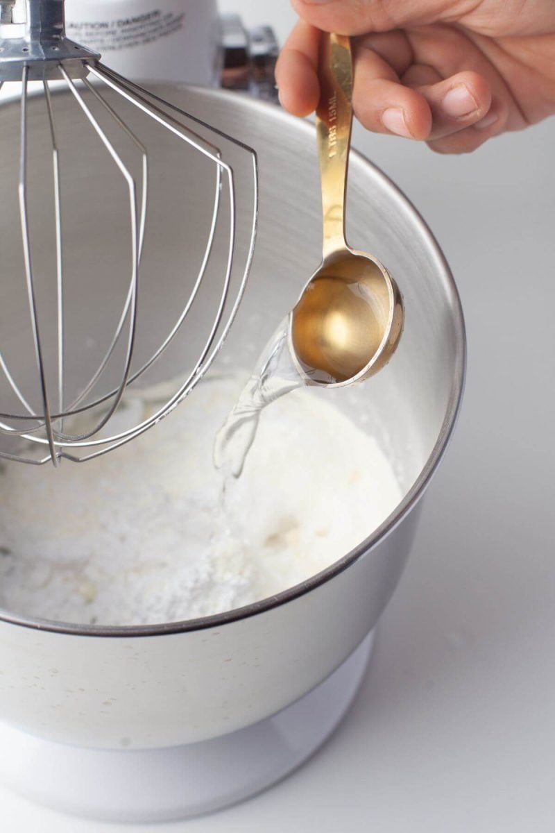 Someone's hand is pouring spoon of vanilla into mixing bowl.