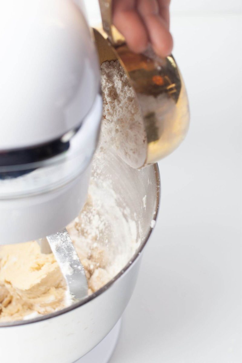 A hand pours flour mixture into butter mixture in stand up mixer bowl.