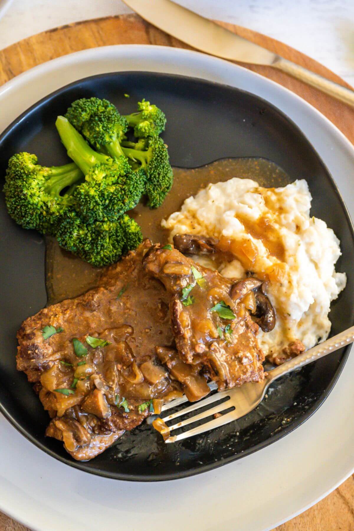 A fork lies next to cube steak, mashed potatoes, gravy and broccoli on plate.