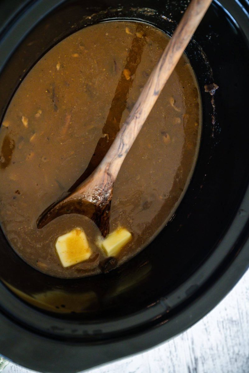 A wooden spoon and chunk of butter rest in crockpot with gravy.