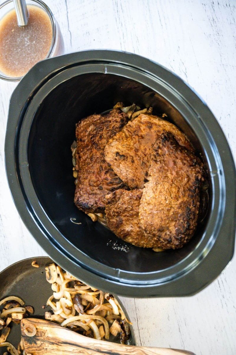 Seared cube steaks rest in crockpot next to plate of onions.