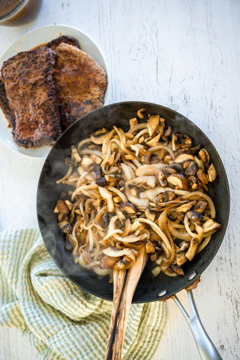 A wooden spoon dips into skillet full of onions and mushrooms.