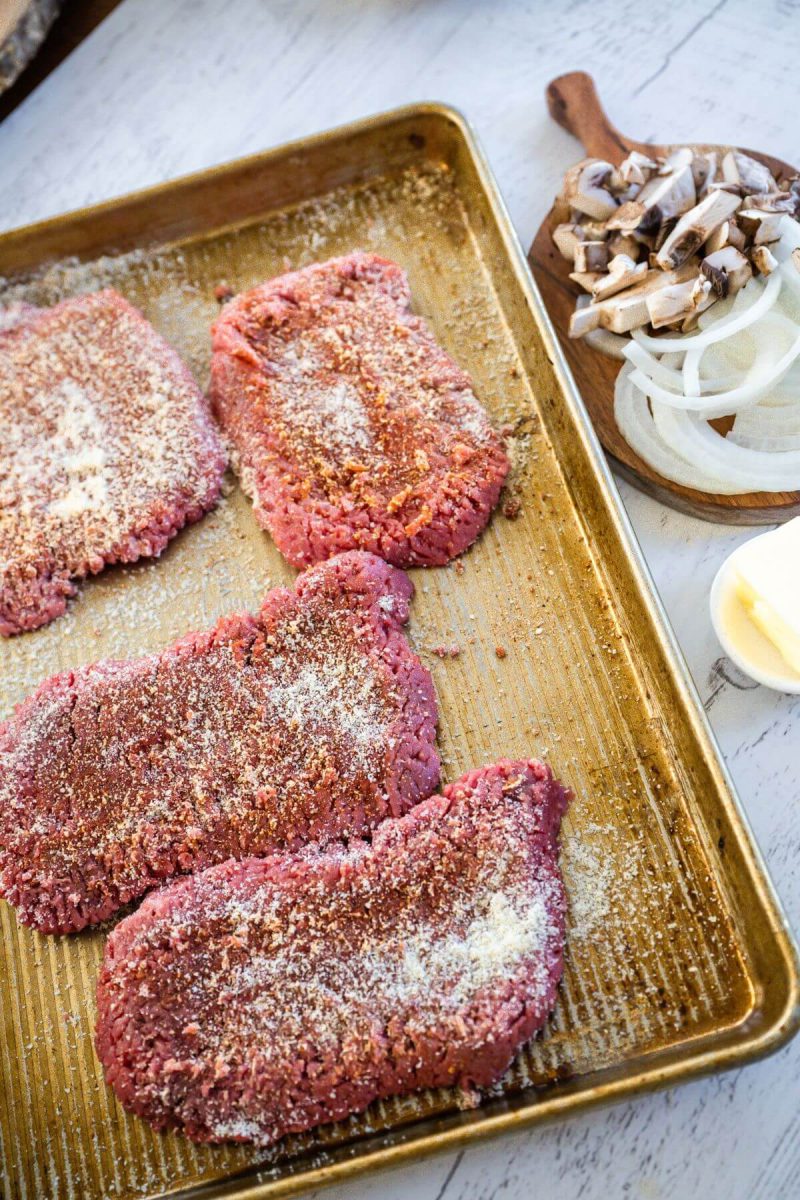 Raw onion and mushrooms sit by raw, seasoned cube steaks with flour sprinkled on both sides.  