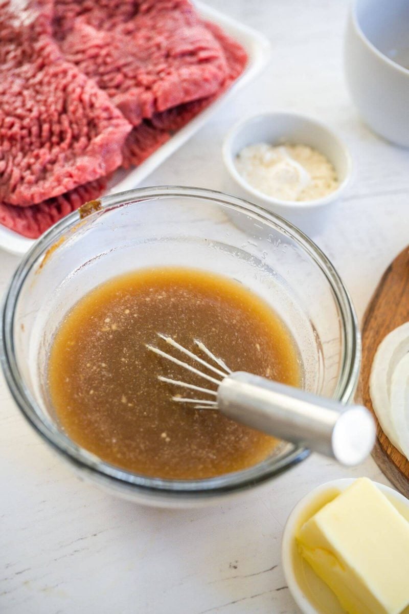 A small bowl of liquids and a whisk rest next to raw steaks.