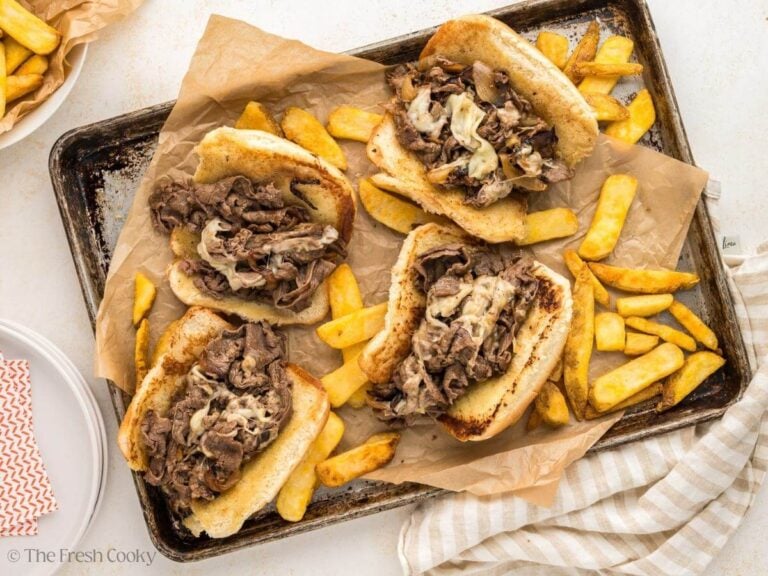Pan filled with 4 Philly Cheese steak sandwiches with french fries sprinkled around.