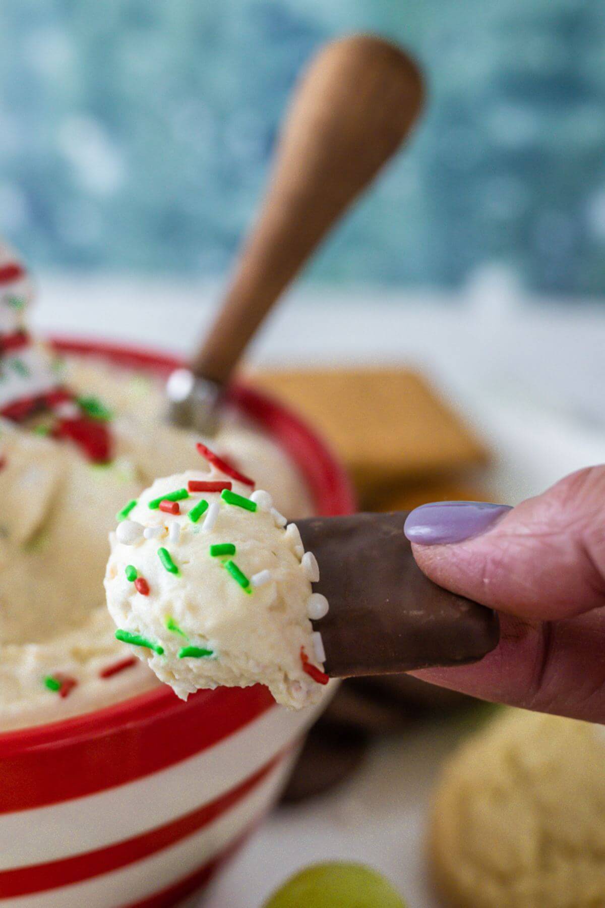 A hand is holding a chocolate graham cracker with Christmas tree cake dip on half of it from the bowl.