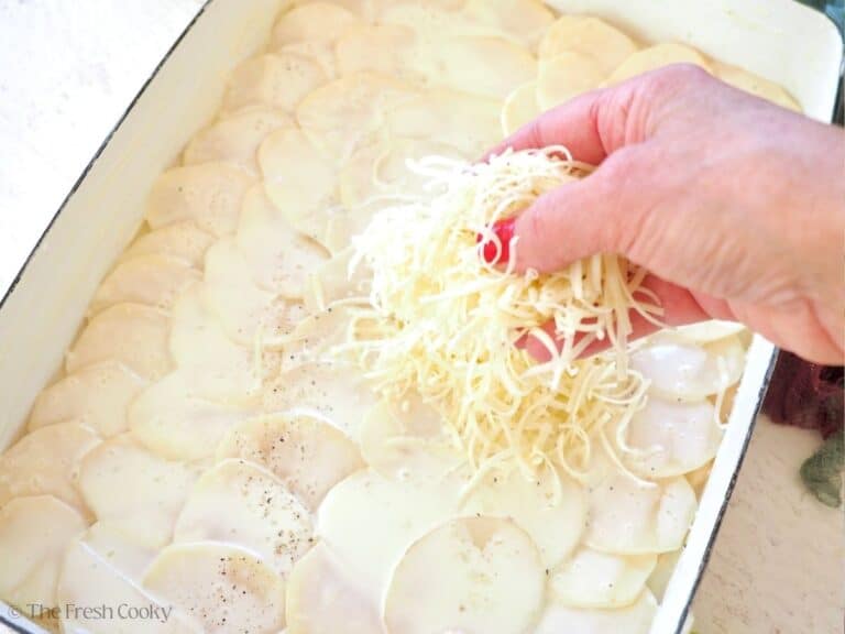 Hand adding a layer of shredded gruyere cheese on top of potatoes.