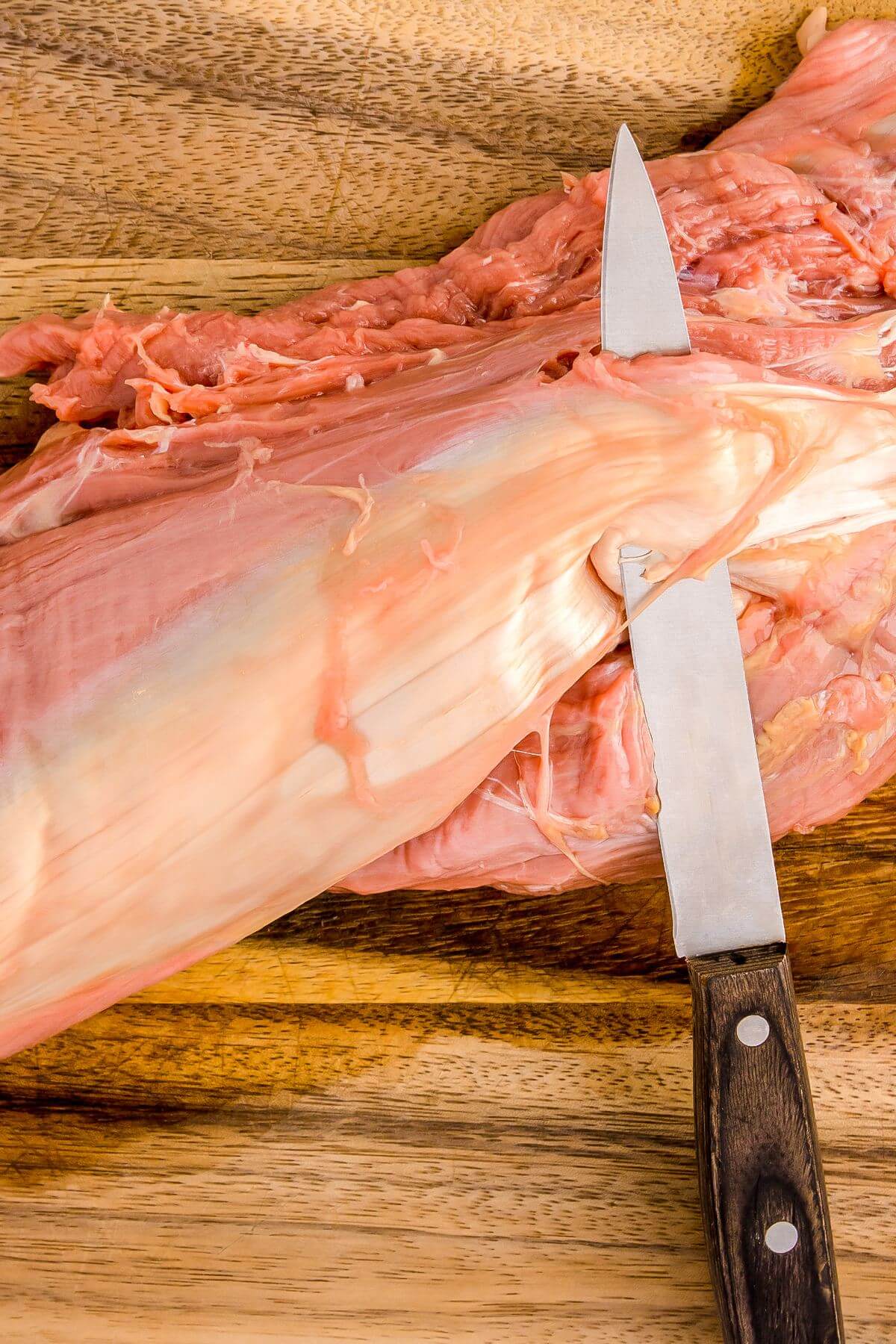 A sharp knife is running under the silver of the raw tenderloin for slicing.