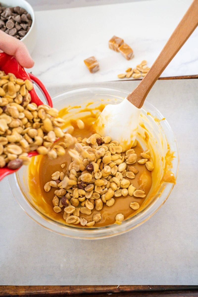 A hand pours peanuts into the caramel mixture in a glass bowl with spatula.