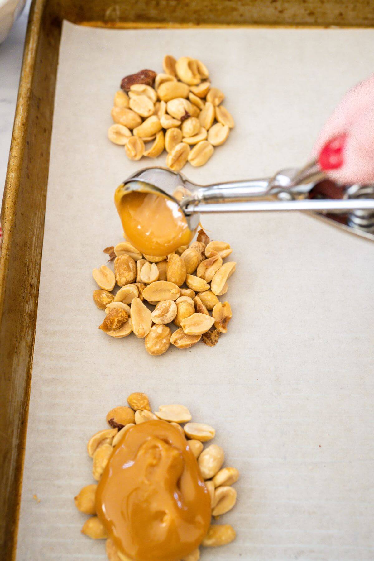 A cookie scoop pours caramel mix on top of peanuts on a baking sheet.