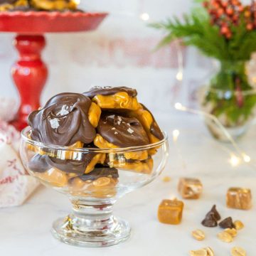 A glass serving dish holds peanut clusters and sits next to holly, caramels, and chocolate chips.