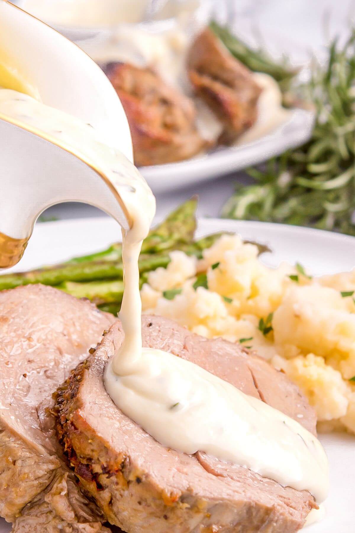A sauce serving holder pours white sauce onto meat dish next to mashed potatoes and asparagus.