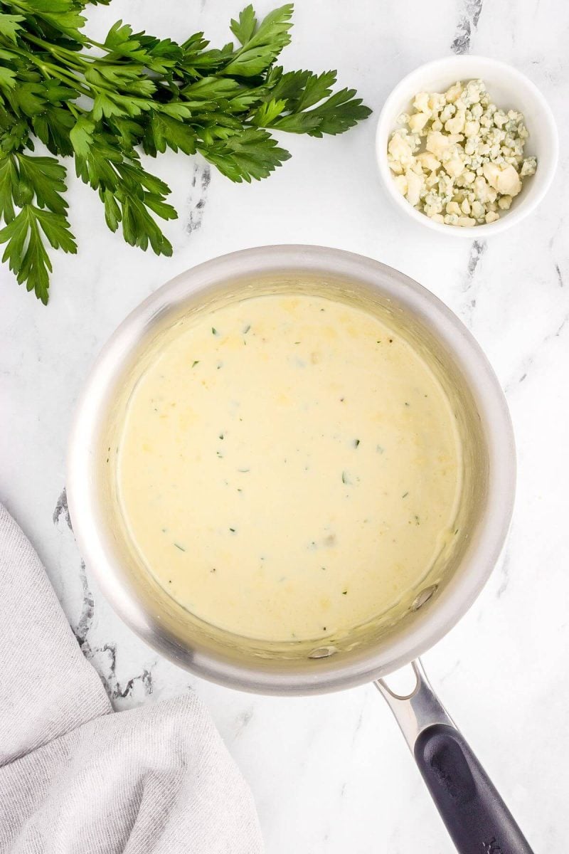 White sauce in pan shows bits of green in it, with parsley and cheese behind pan.