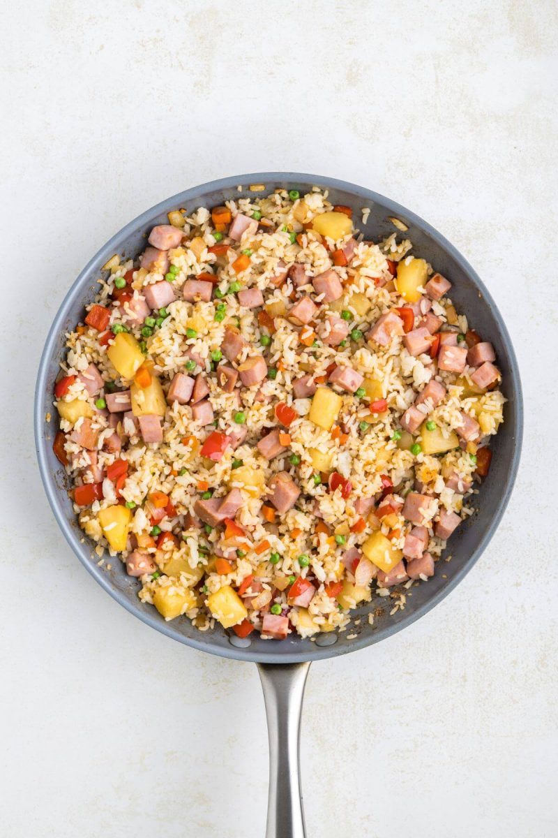 A saucepan is full of rice, ham, pineapple, peppers, and onions.