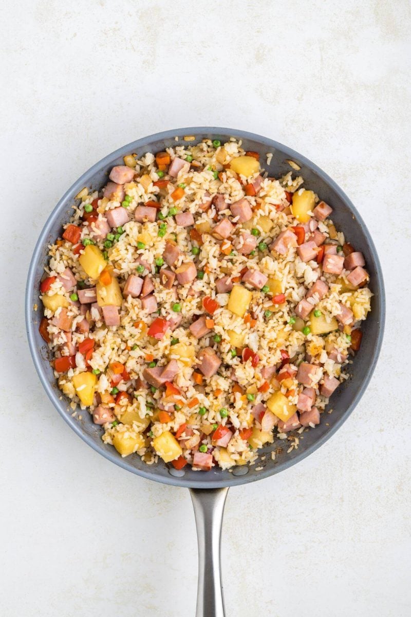 A saucepan is full of rice, ham, pineapple, peppers, and onions.