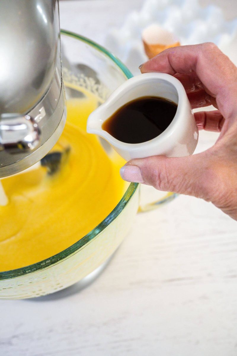 A hand holds a small spouted cup with vanilla extract to pour into mixing bowl.