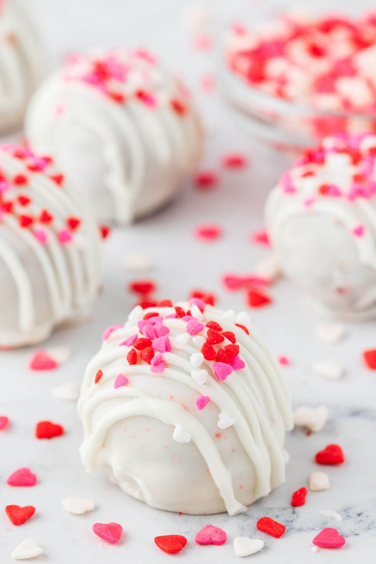 White iced cake ball has pink and red heart sprinkles on top and surrounding table.