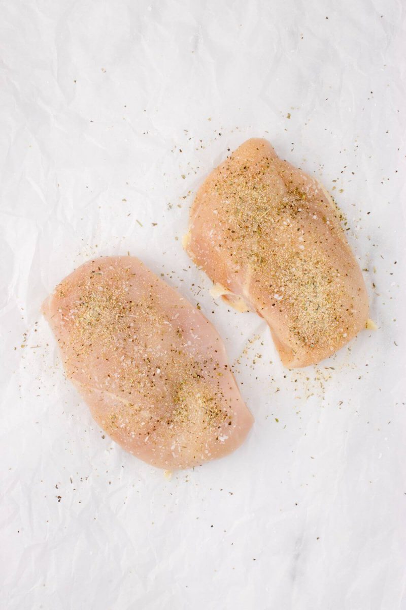 Spices are sprinkled over raw chicken breasts on table.