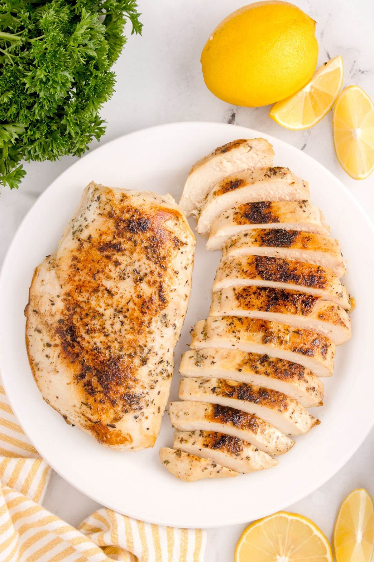 A plate full of both chicken breasts, one whole and one sliced, is next to greens and lemons on table.
