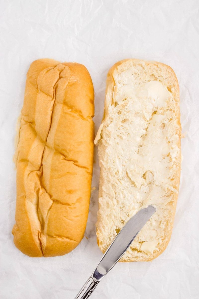 A knife is spreading butter on a bun.