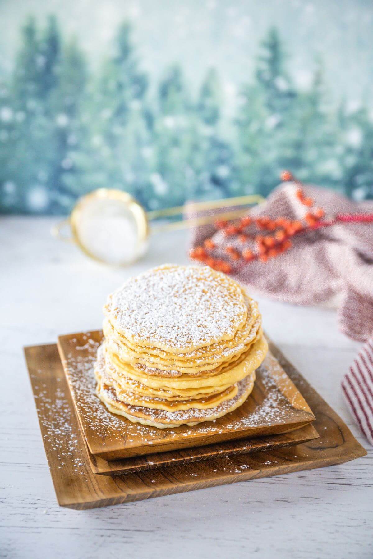 Tall stack of cookies sit on tiered plates in front of wintery scene with trees and red berries.