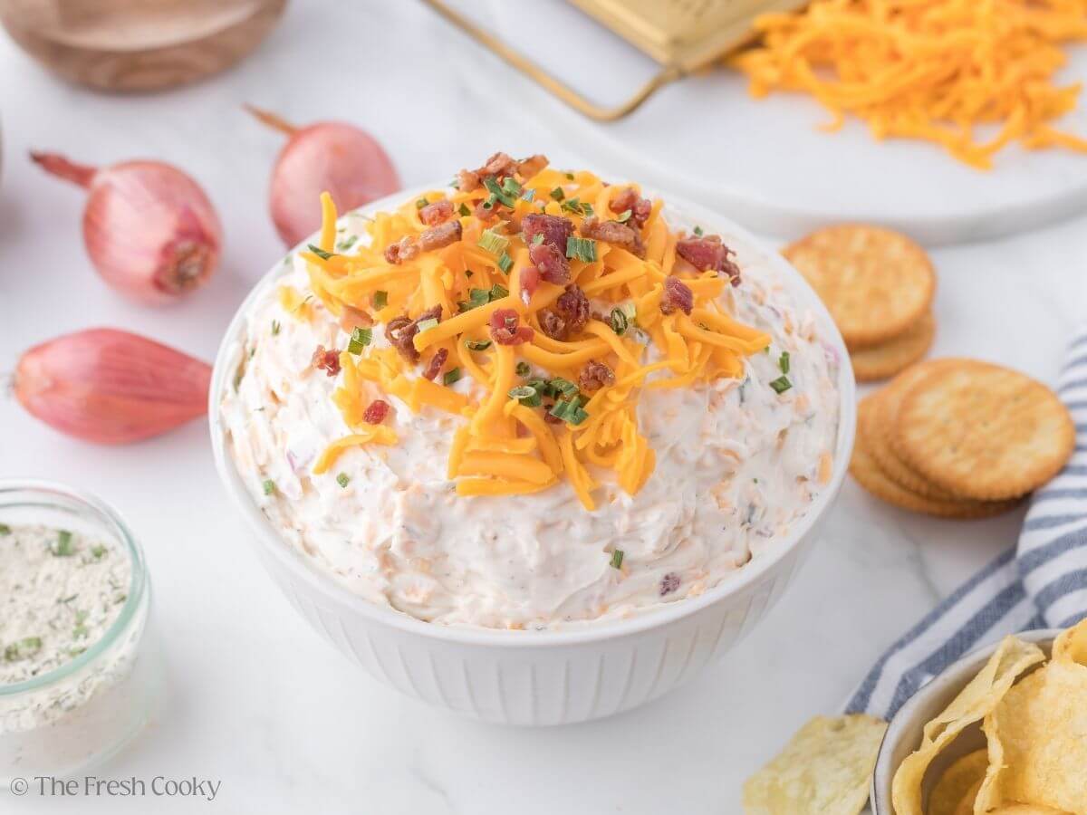 A white bowl filled with creamy, cheesy, crack dip, topped with extra shredded cheddar, bacon bits and green onions.
