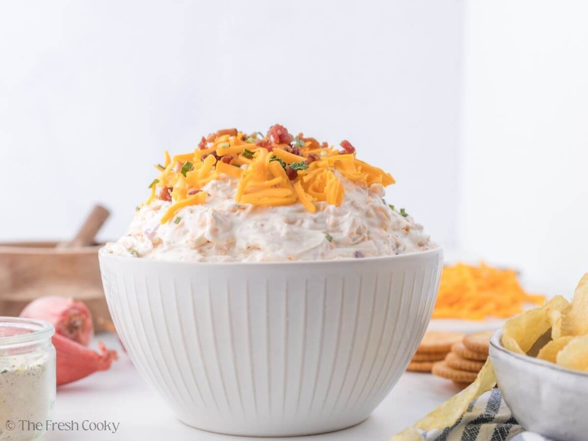 Looking at a side-view of mounded, crack dip, creamy and cheesy, topped with extra shredded cheddar cheese, bacon bits and green onions.