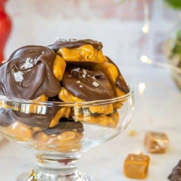 A glass serving dish holds peanut clusters and sits next to holly, caramels, and chocolate chips.