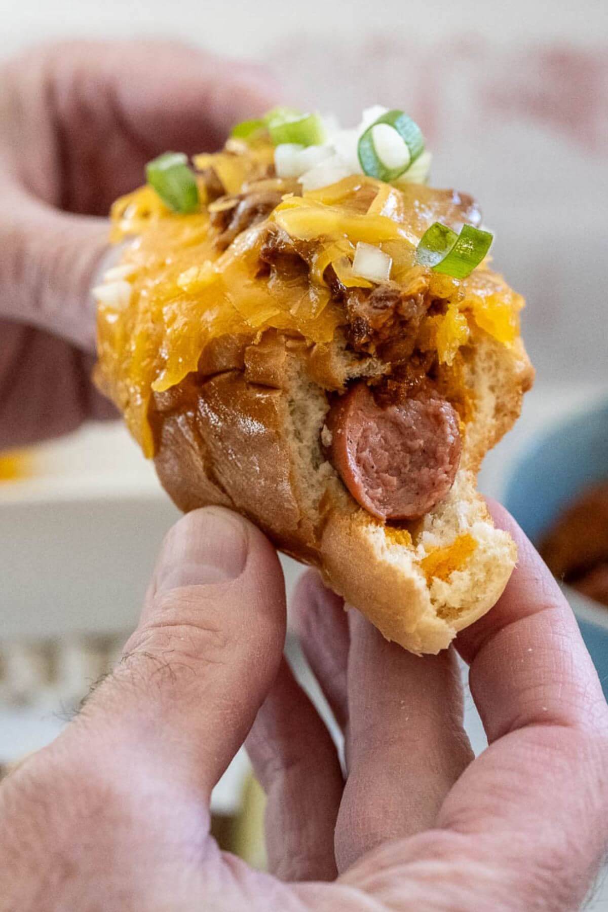 A half eaten chili dog is held between two hands showing all the layers of toppings.