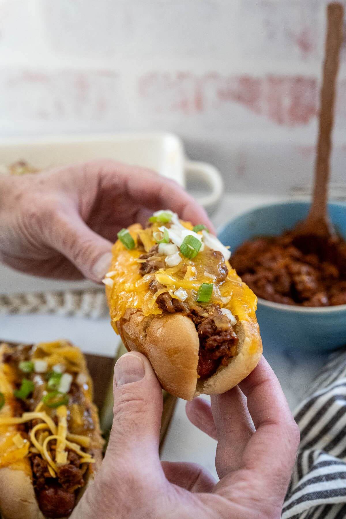 A chili dog is held by two hands above the full casserole dish.