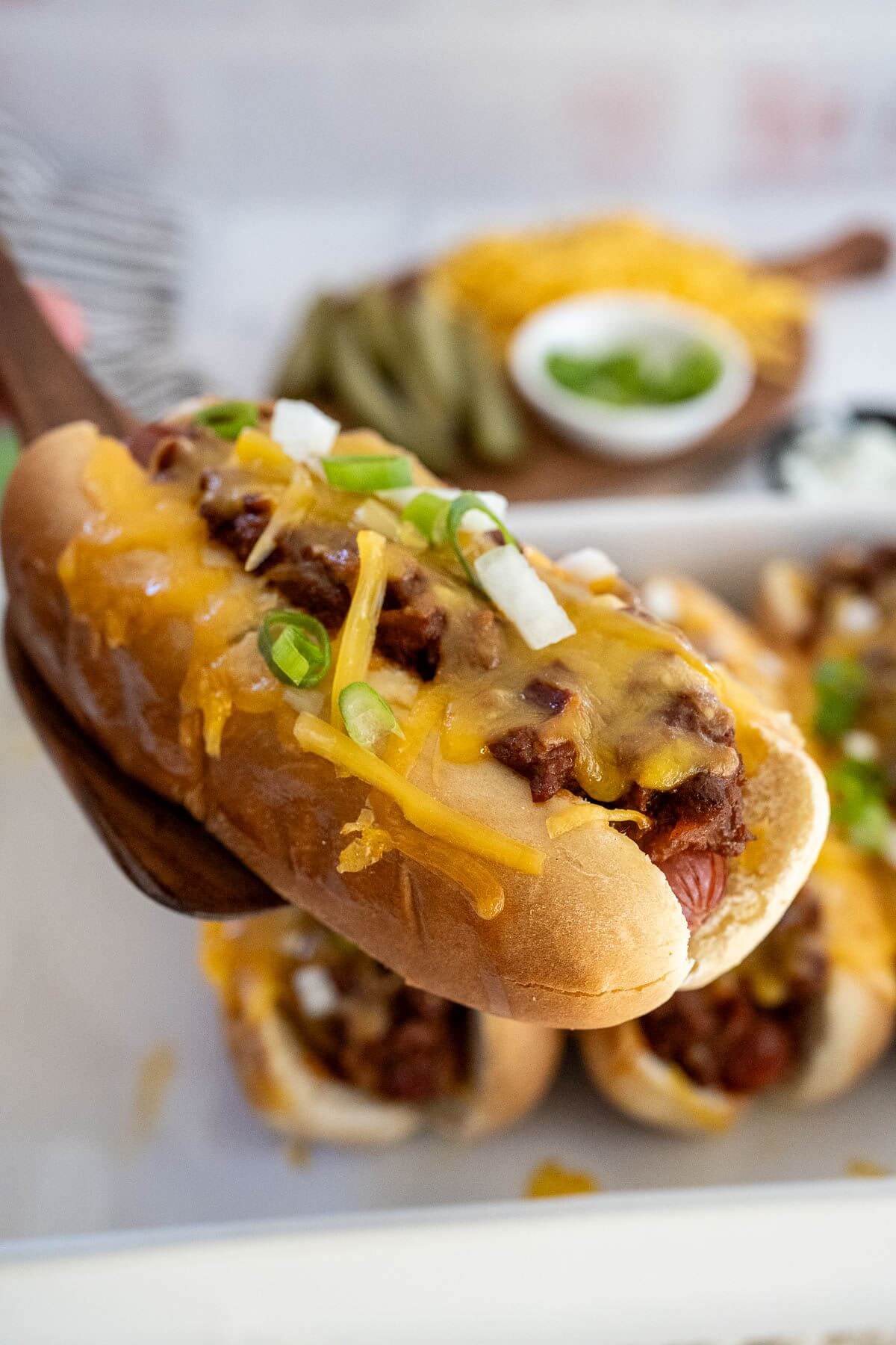A melty, cheesy chili dog with white and green onions is lifted from the baking dish by a wooden spatula.