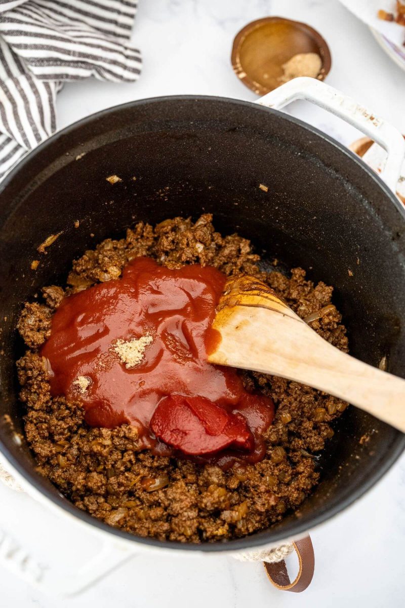A wooden spatula stirs tomato sauce into ground beef mix.