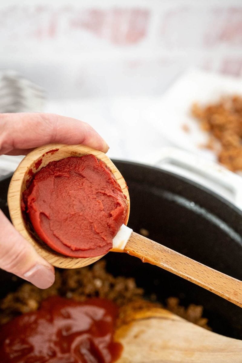A spatula scoops out tomato paste from a bowl a hand is holding.