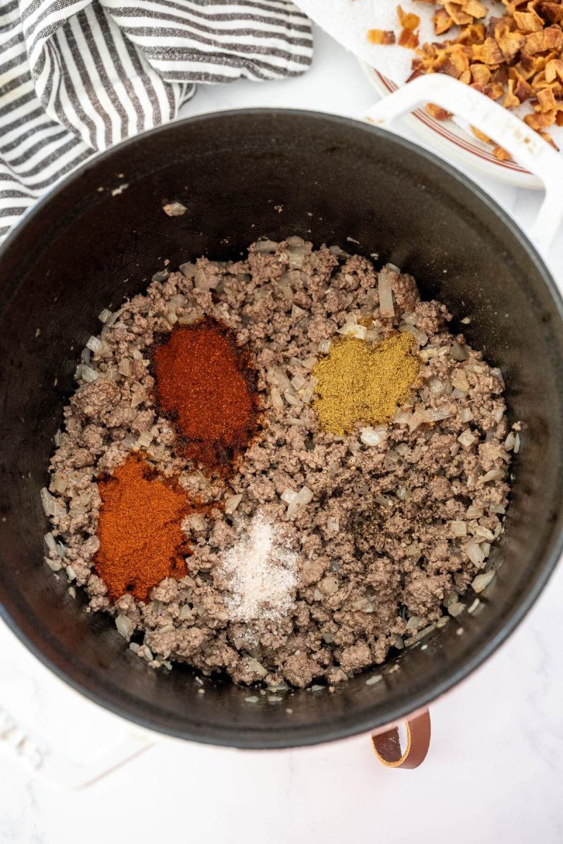 Cumin, smoked paprika,, salt and chili powder are on top of cooked ground beef.