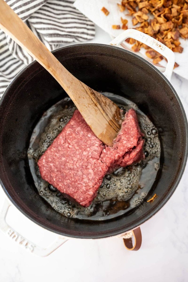 Raw ground beef is in large pot being broken and stirred by wooden spatula.