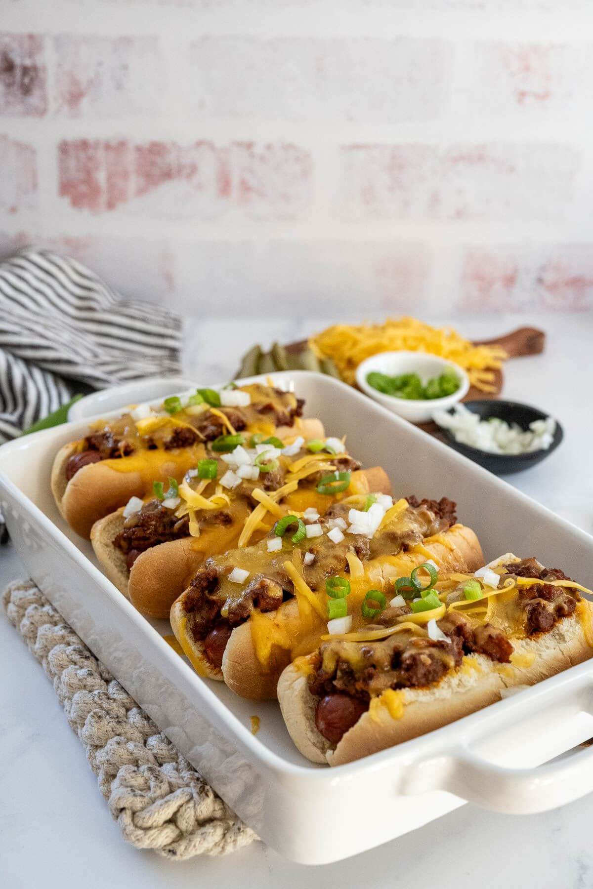 Small bowls of extra toppings are next to the big baking dish filled with cooked chili dogs.